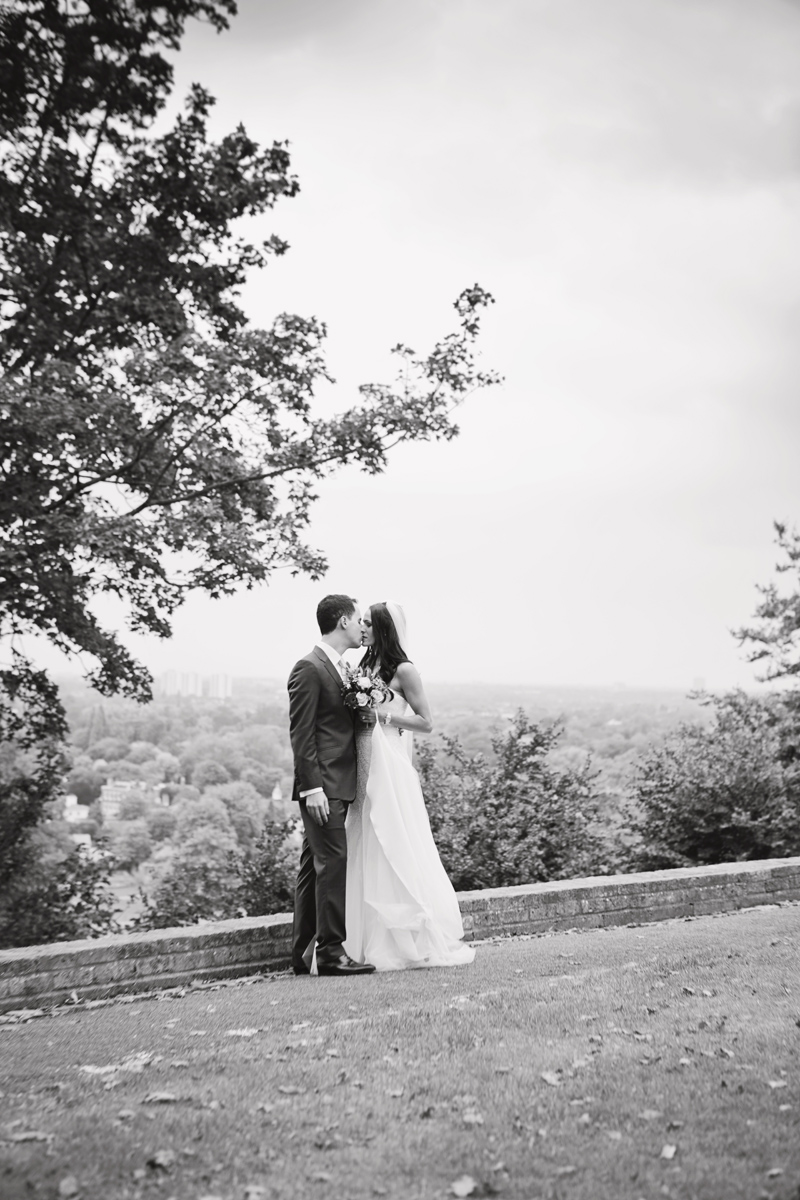 A newlywed couple kiss with views of the Thames Valley behind them in Richmond Park