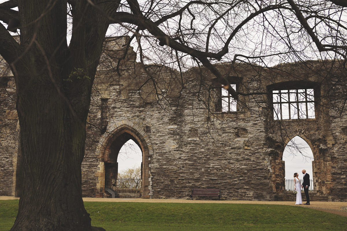 A wide shot of a bride and groom facing each other in the grounds of Newark Castle with a tree in the foreground