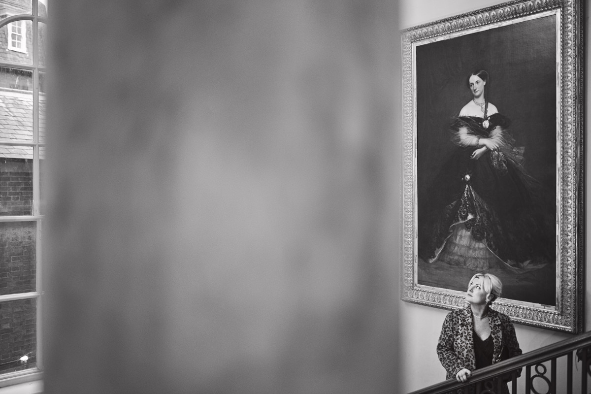 A black & white image of a women standing at the top of a large staircase admiring the old paintings in a grand hall
