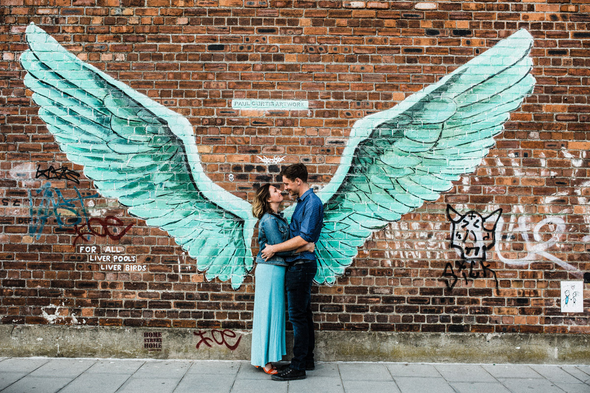 A young couple embrace in front of graffiti depicting a pair of wings in Liverpool