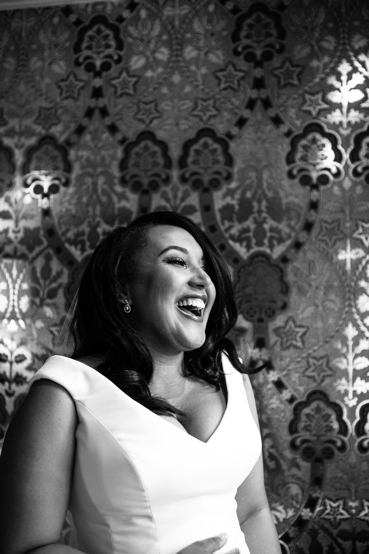 A black and white shot of a laughing bride stood in front of patterned wallpaper