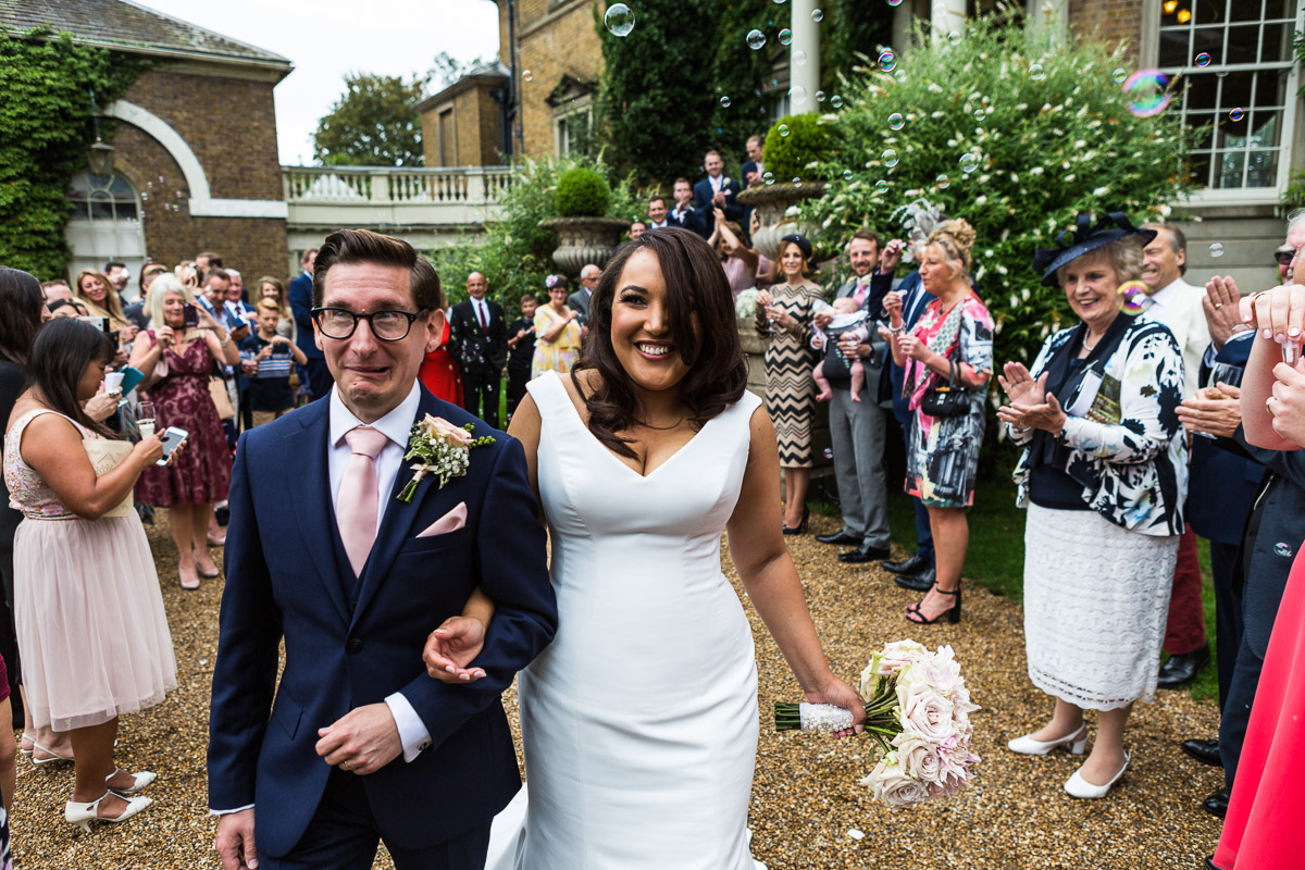 A groom pulls a funny face as newlyweds smile surrounded by their guests at Hampton Court House