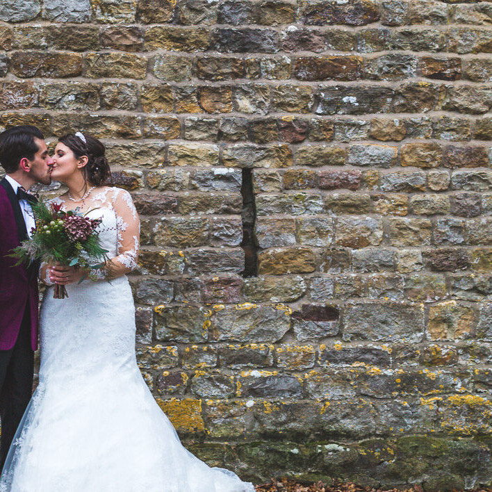 A wedding couple kiss while stood in front of a rustic wall during a winter wedding at The Out Barn in Clough Bottom
