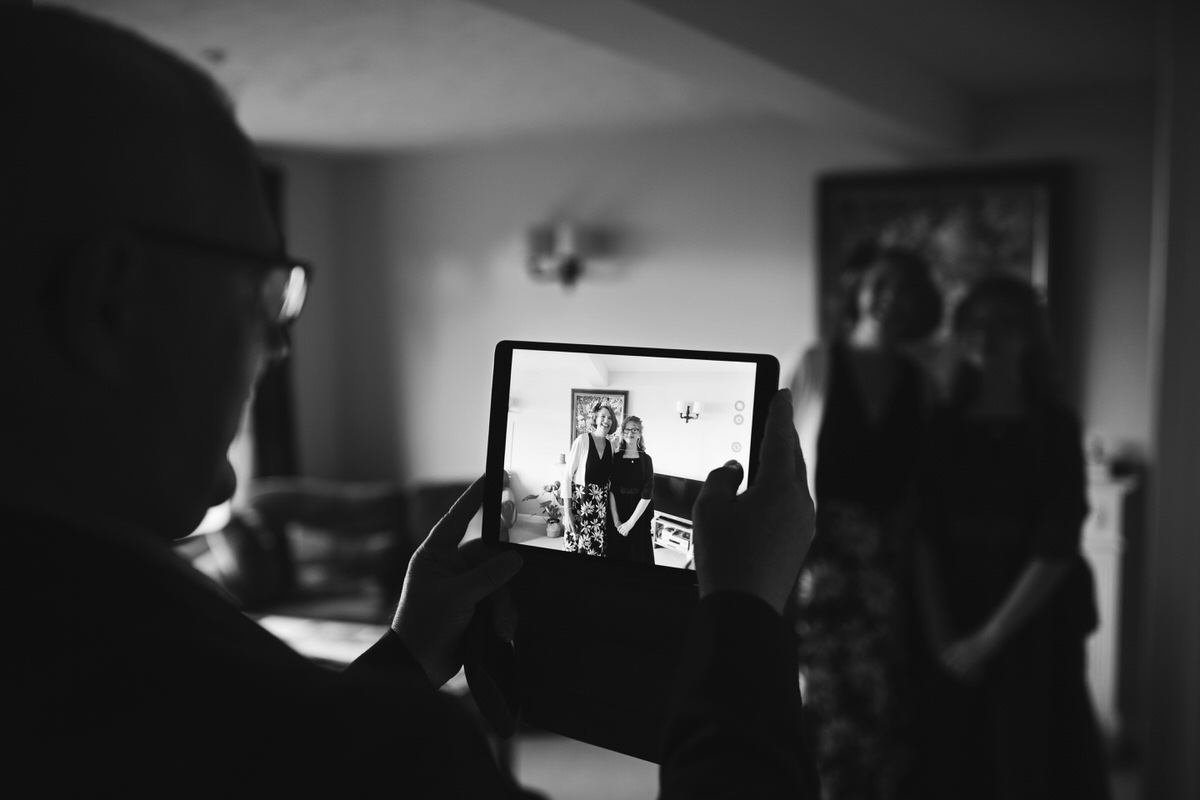 A black and white shot of a man holding an iPad taking a photo of family in a house