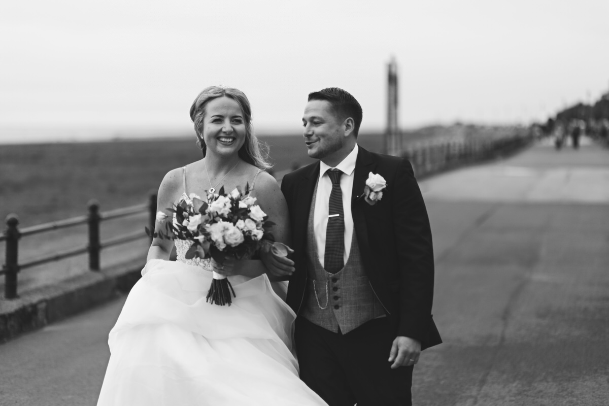 A wedding couple walking along the promenade of a seafront, smiling