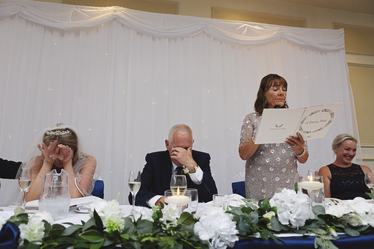 A bride and best man cover their faces in embarrassment during the mother of the bride's speech