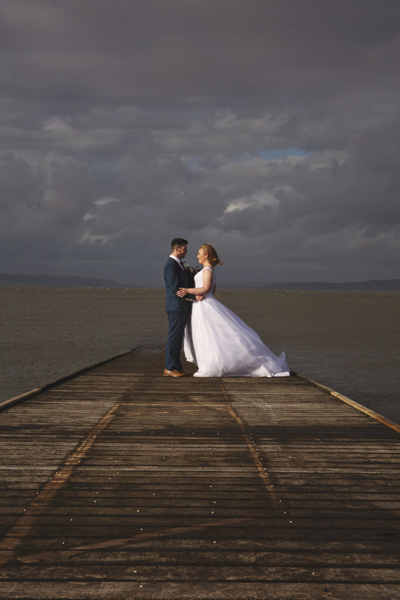 A bride and groom face each other on the end of a jetty by the sea